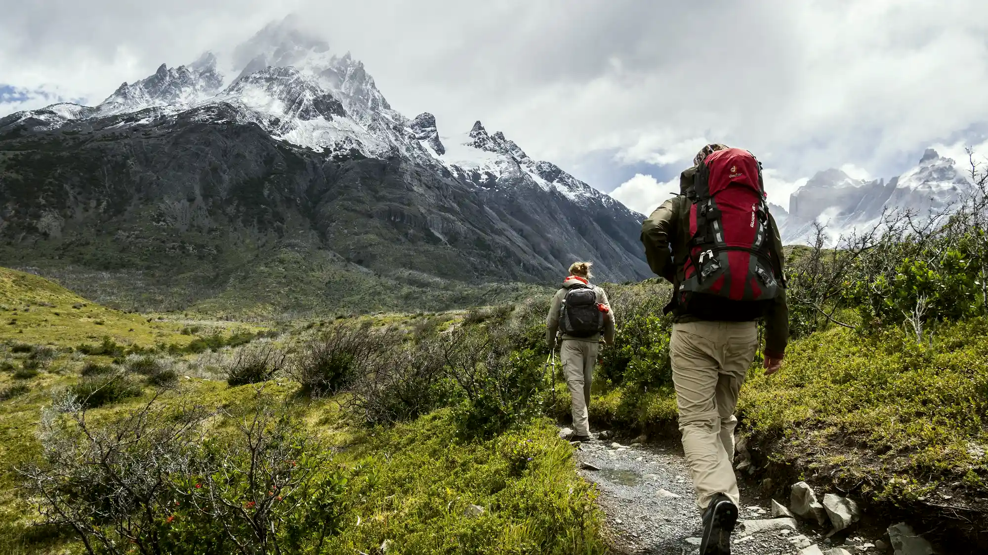 Two hikers on a mountain trail below snow-capped peaks, seen from behind.