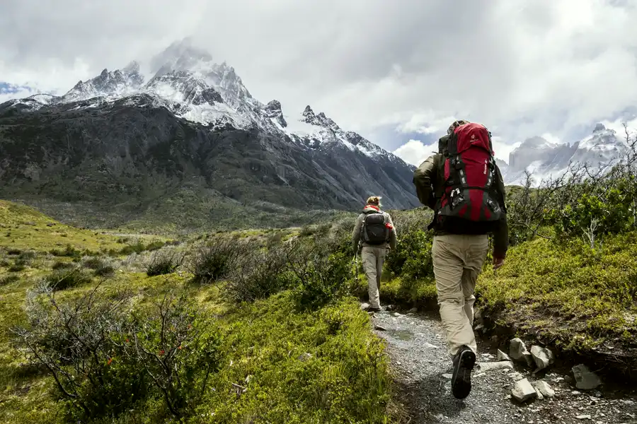Two hikers on a mountain trail below snow-capped peaks, seen from behind.